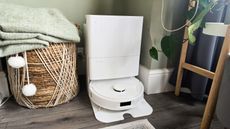 A white Roborock Qrevo S Vacuum in its base with a woven basket and a plant stand either side of it