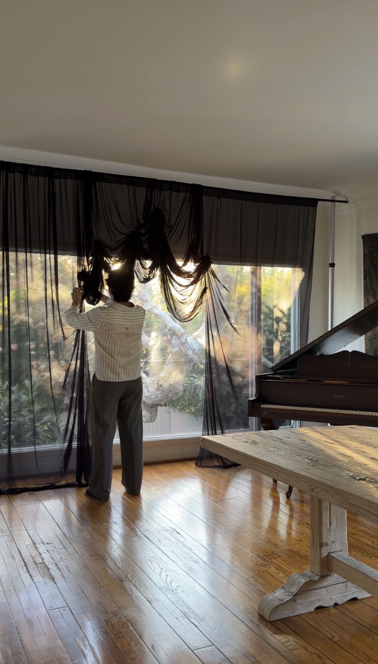 A man ruching a black sheer curtain panel in front of a window to expose the trees outside next to a black grand piano in front of a wooden dining table with nothing on it
