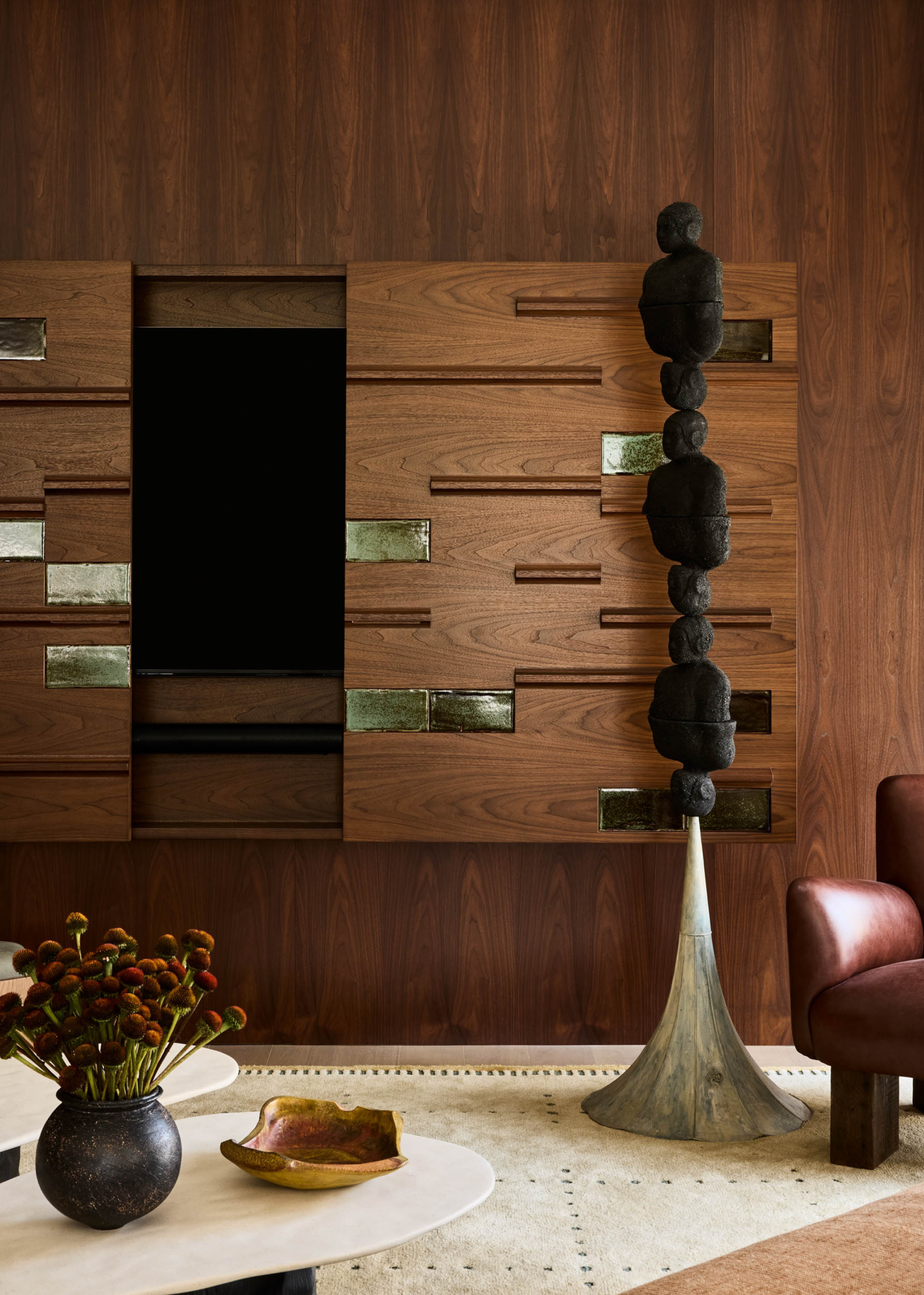 A wood living room with a stacked statue, a leather armchair, a marble coffee table with a vase of flowers, a ceramic bowl, and a TV concealed by wooden doors with green gem accents