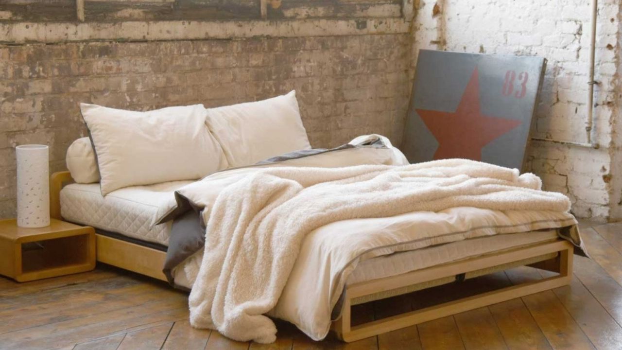 A futon mattress with beige bedding against an exposed brick wall.