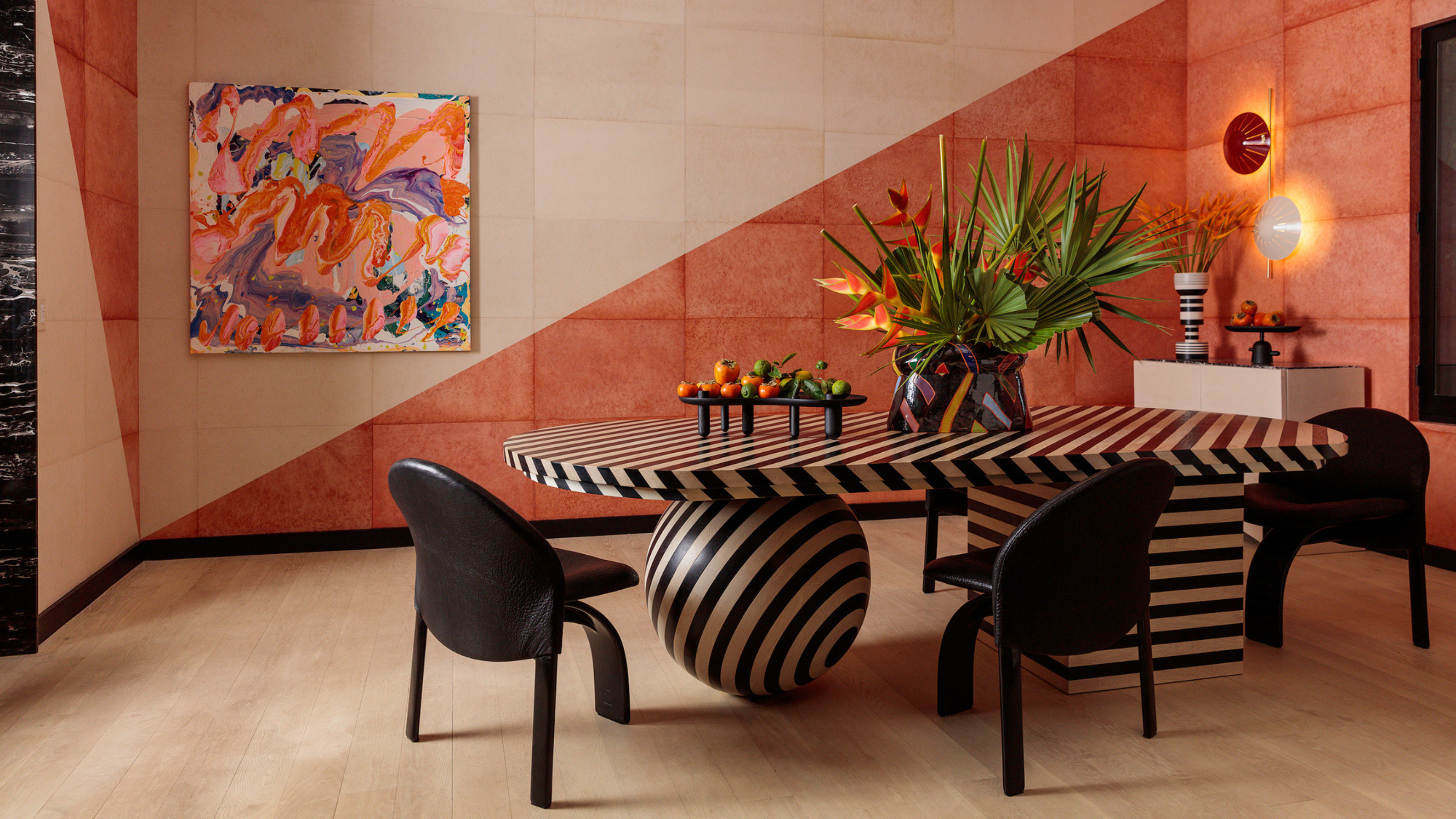 A dining room with a bold striped dining table with one ball leg and one square, with black dining chairs and a wall painted half pale pink and half salmon pink on either side of a diagonal