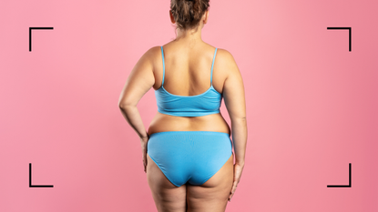 Plus size woman in blue underwear with her back to the camera. She is standing against a pink background