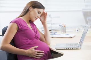 A pregnant woman sits at her desk holding her head