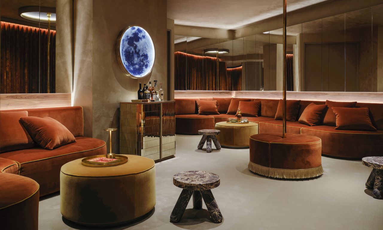 Club room with custom rust-coloured curved sofas, mirrored panels on the walls, mirrored bar cabinet, light sconce and red marble side tables