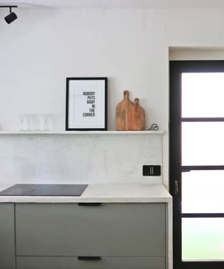 A kitchen with microcemented walls, a wall shelf with wall art and chopping boards on it, a white countertop with a gray cabinet, and a black PVC door