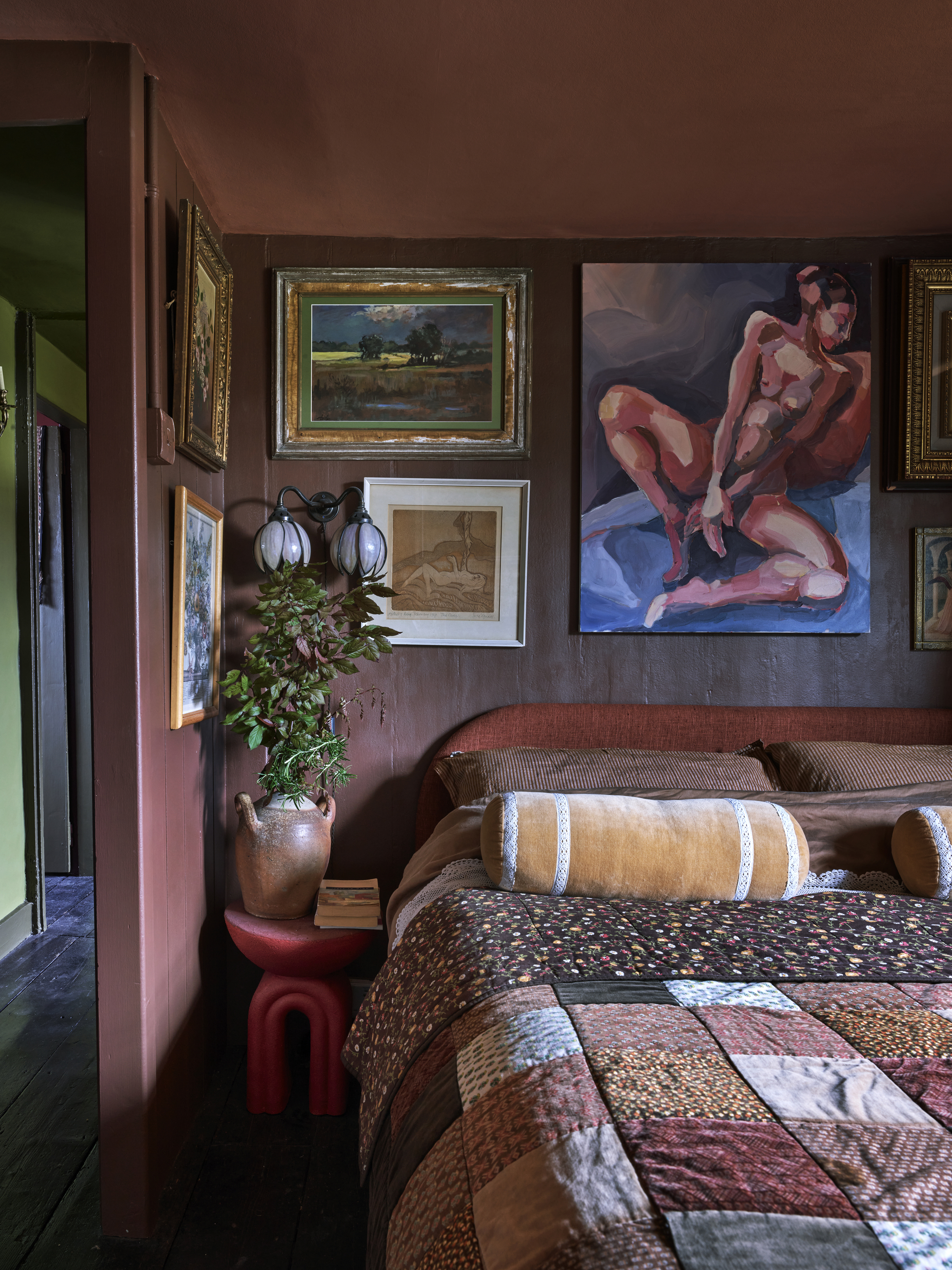 bedroom with dark red walls, gallery wall of artwork, red side table with terracotta vase with branches and patchwork quilt on the bed