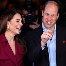 Kate Middleton wearing a red dress and laughing standing next to Prince William wearing a suit jacket and blue dress shirt and pointing and laughing at someone