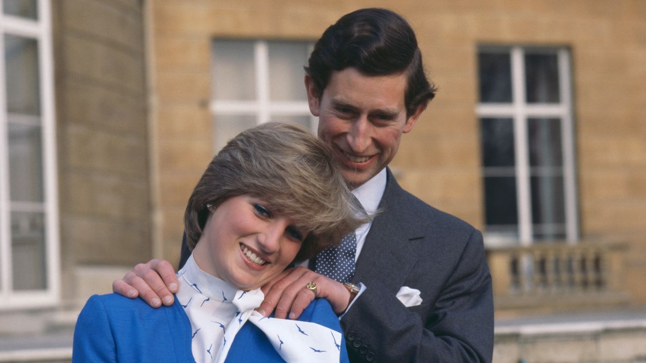 Prince Charles wearing a gray suit laughing and posing with his hands on Princess Diana&#039;s shoulders in front of a staircase in their engagement photos 