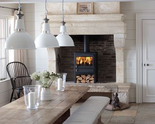 Stove in large natural stone fireplace in dining room, log storage beneath stove, large wooden dining table white paneled walls, white painted door, three low hanging pendants over table