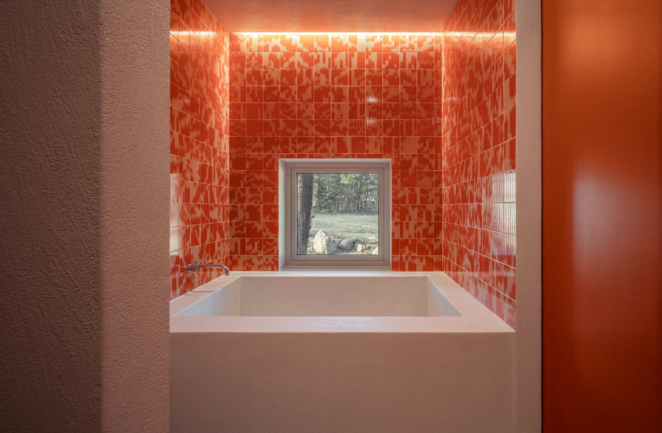 Pale pink built-in bath surrounded by glossy orange patterned tiles, with concealed ceiling lighting and a small window overlooking trees.