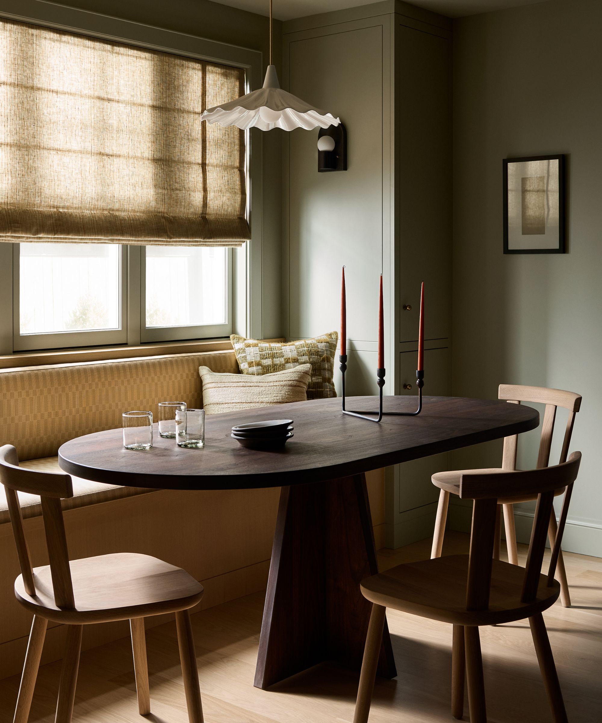 a cozy breakfast dining nook with large window, linen blind, a dark wooden table and a banquette seat with oak chairs and a fluted pendant overhead