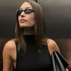 Hailey Bieber in an elevator wearing tiny sunglasses, a sleeveless turtleneck, a Bottega Veneta Falcon clutch, and a pencil skirt with two belts.