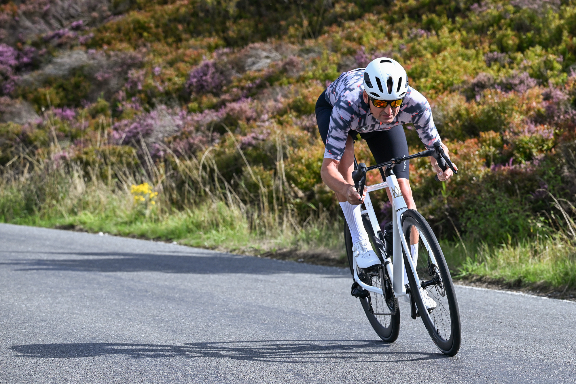 a white road bike being ridden downhill by a man wearing white shoes, white helmet, black shorts and a patterned grey jersey with purple heather in the background