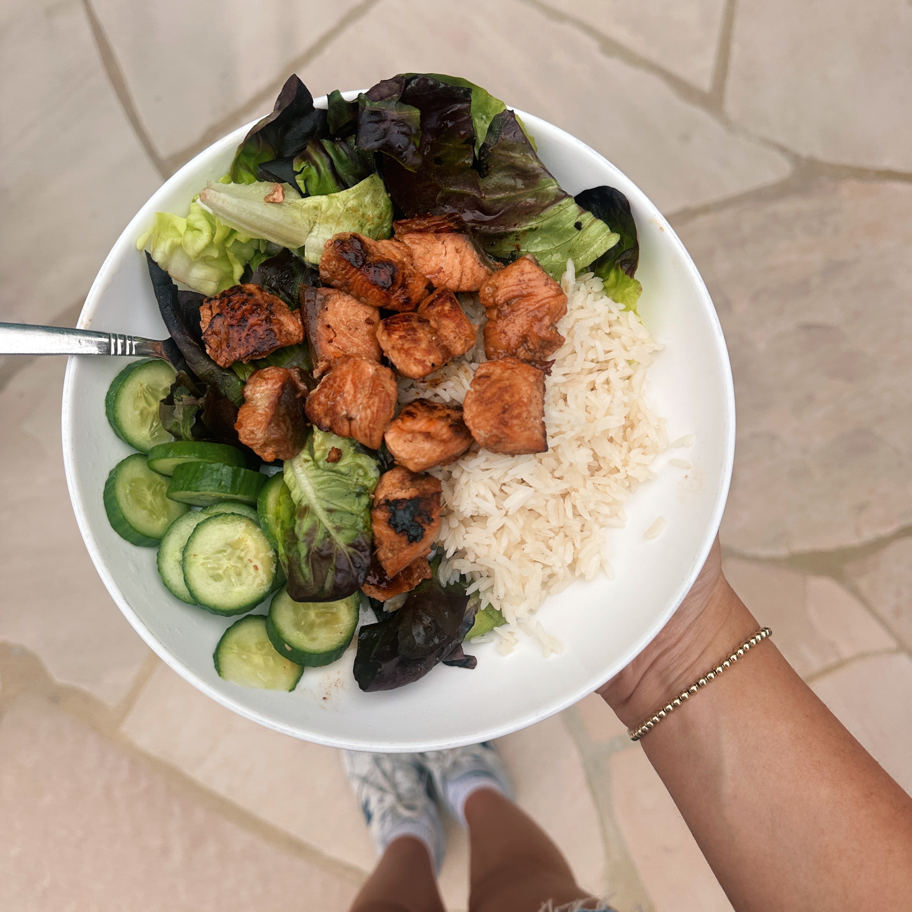 Hand holds bowl of rice, salmon chunks, cucumber and lettuce