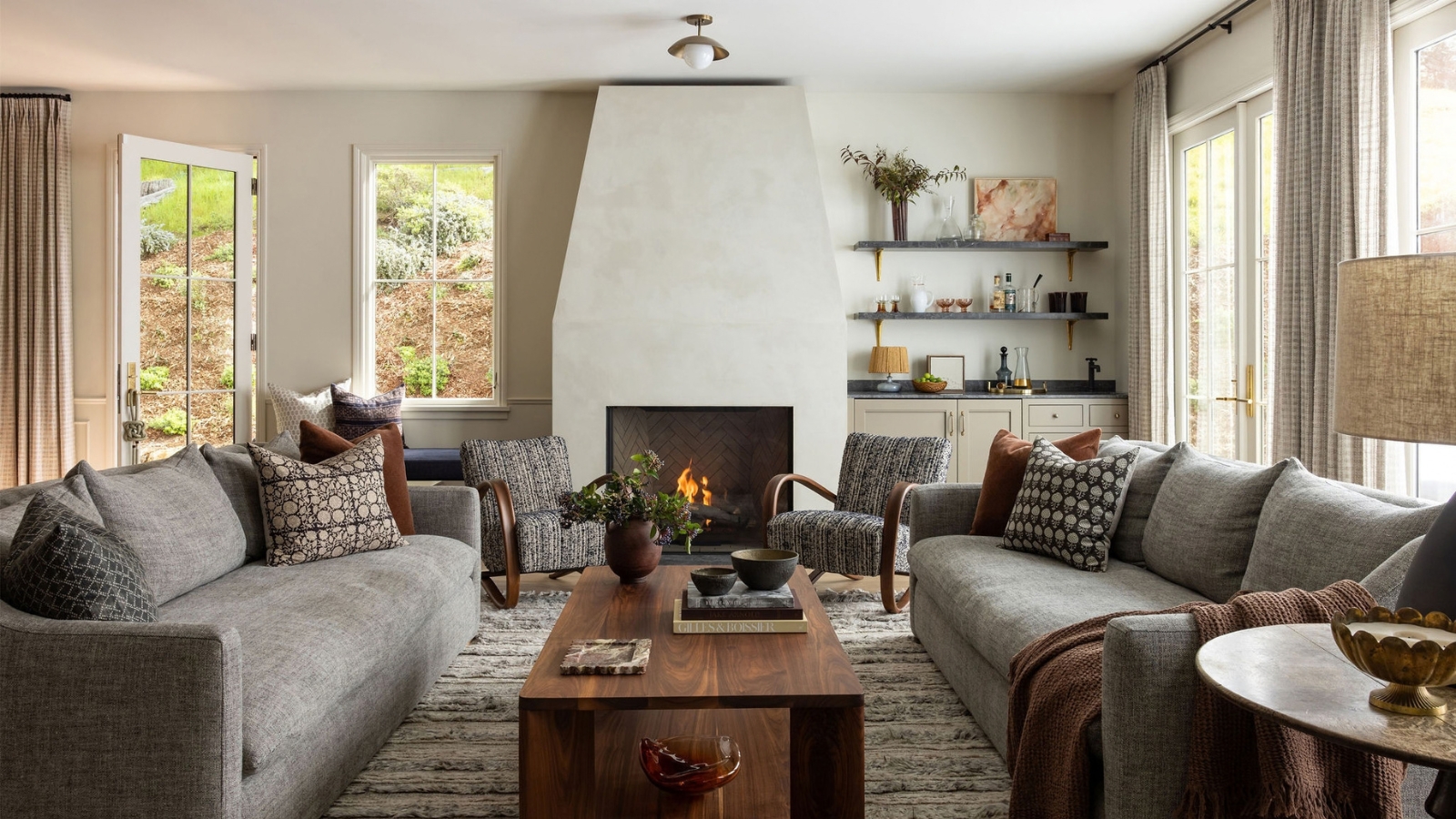 Living room with facing gray sofas, patterned pillows, a wood coffee table, curved armchairs and a fireplace beside a built-in bar.