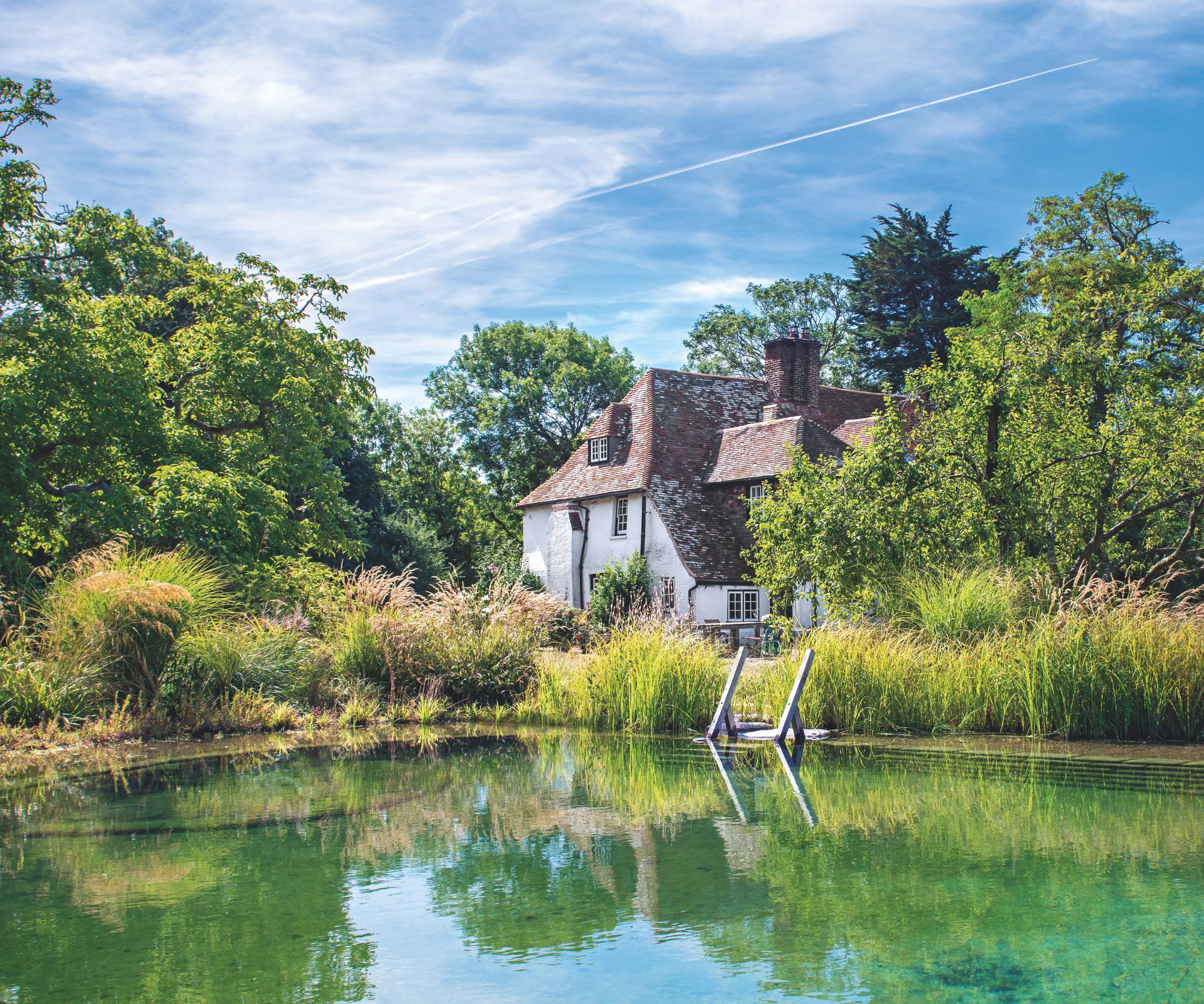 Historic country cottage with a natural swimming pool in front, surrounded by wild grasses and mature trees under a bright blue sky.