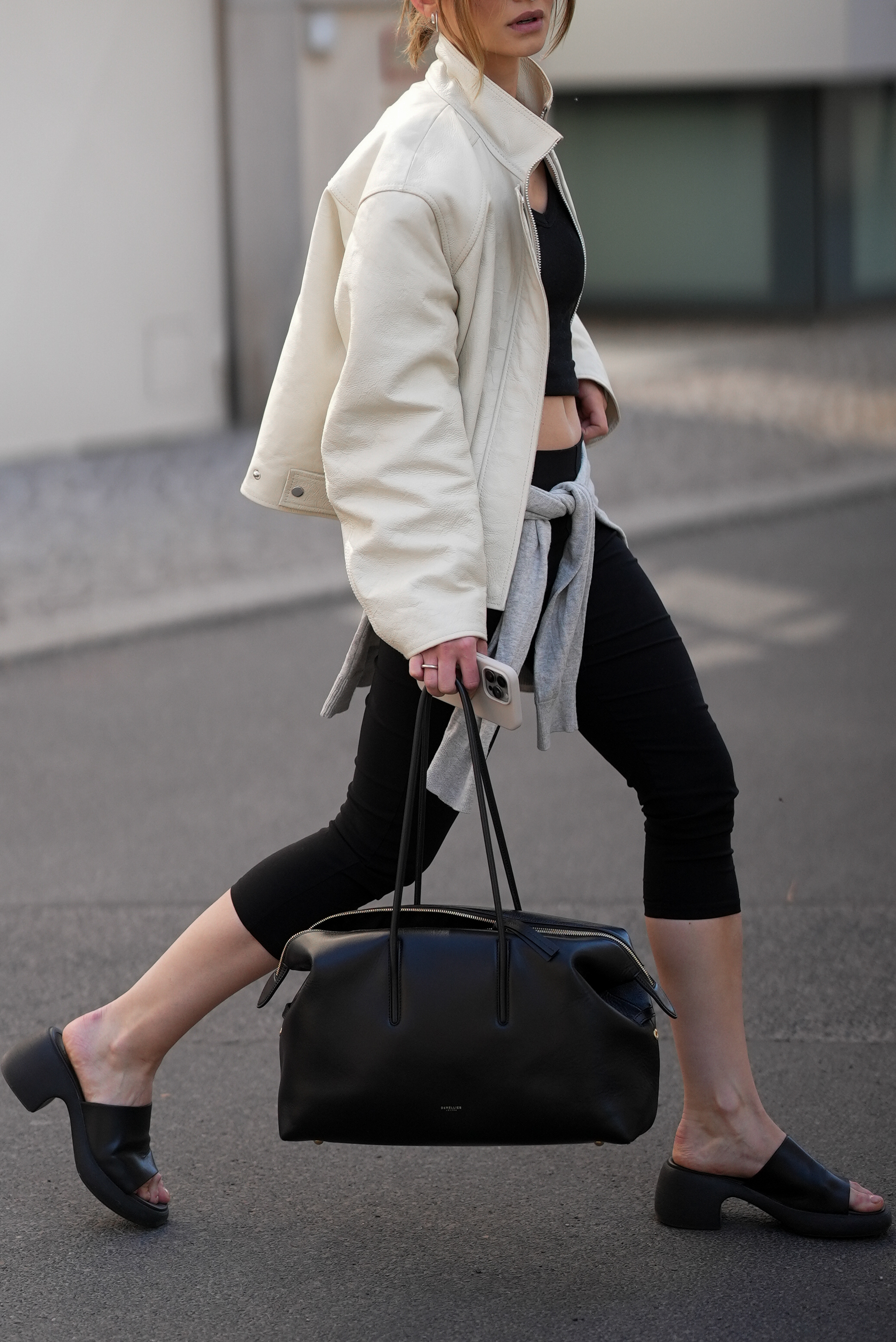 BERLIN, GERMANY - MAY 3: Sophia Geiss is seen wearing an off-white oversized harrington-style jacket with a stand collar, zip closure, and dropped shoulders from Samsoe SamSoe; a black cropped V-neck top with a fitted silhouette from Weekday; black cropped leggings in a stretchy fabric, ending below the knee from Mango; a light grey pullover tied around the waist from Munthe; black chunky platform mule sandals with a wide single strap, open toe, and a thick block heel from Camper; a natural pedicure; "The Stockholm" large structured black leather tote bag with gold hardware and double top handles from DeMellier; holding an iPhone from Apple; a nude nail polish manicure; silver and gold mixed jewelry including rings on May 3, 2026 in Berlin, Germany. (Photo by Moritz Scholz/Getty Images)