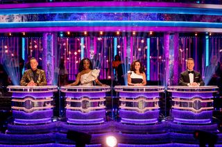 Judges Craig Revel Horwood, Motsi Mabuse, Shirley Ballas, and Anton Du Beke sitting behind their individual desks (divided by transparent screens) in the Strictly studio