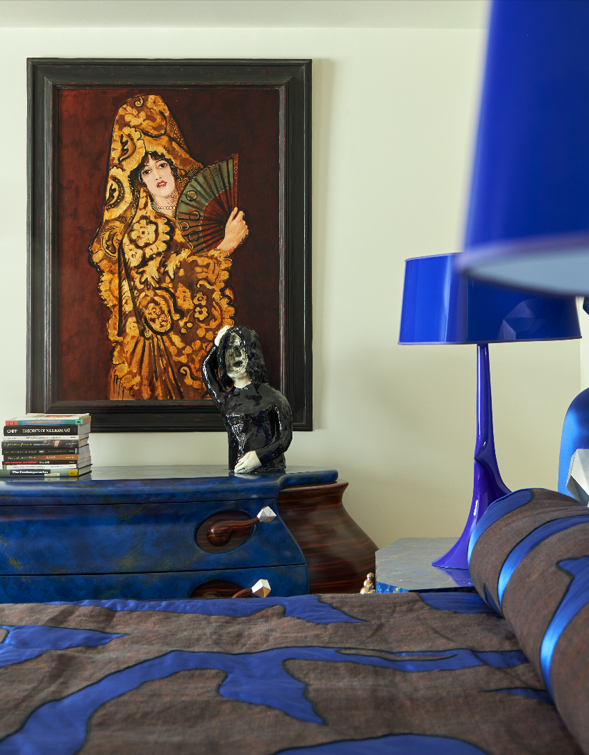 Pauline Karpidas's house interior — a blue and grey bedspread and blue lamps stand in front of a blue dresser with brown handles. A work of art hangs in the background