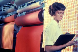 Textile production shot, young dark haired woman in yellow blouse, looking down at a black clipboard that she is holding, rolls of different coloured textiles to the left