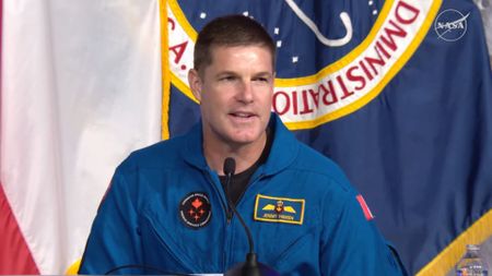 A man with gray hair wearing a blue flight suit with a circular CSA logo answers a question using a microphone on the desk in front of him
