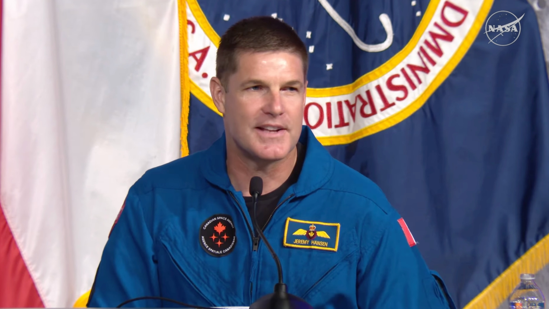 A man with gray hair wearing a blue flight suit with a circular CSA logo answers a question using a microphone on the desk in front of him