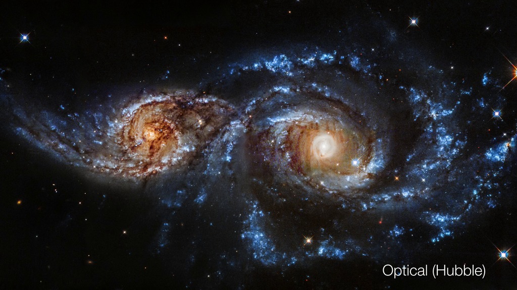 two spiral galaxies colliding with each other. The galaxy on the left appears to be smaller and more brown/orange in color while the one on the right is larger and looks more blue.