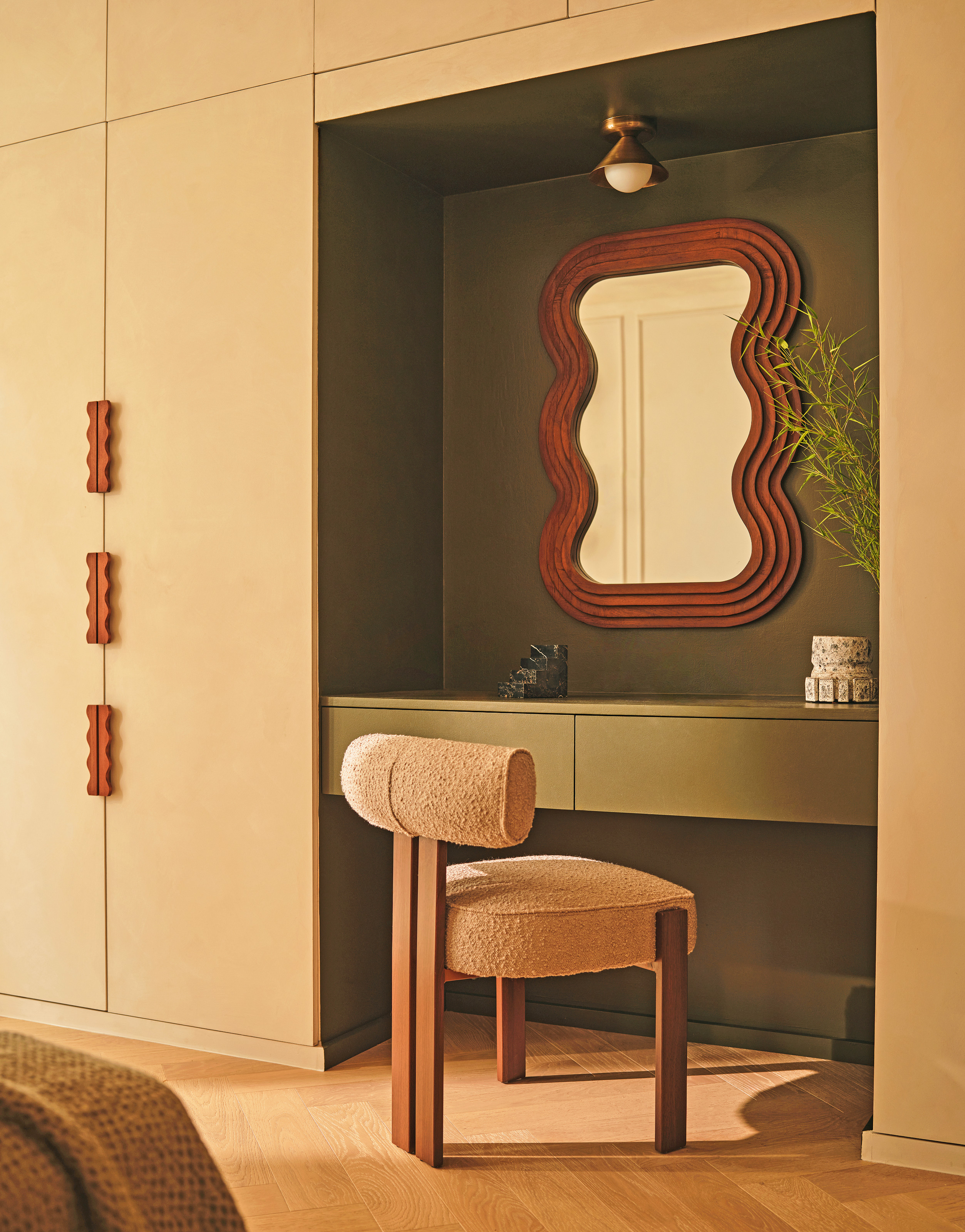 Bedroom corner with built-in wardrobes with wiggle handles, green built-in desk niche, rounded mirror and beige upholstered chair