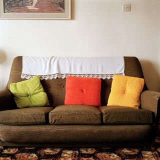 A brown three seater sofa with a white cloth on the headrest, 3 colourful cushions (Green, Red and Yellow), photographed against a white wall with framed wall art and black flowery carpet