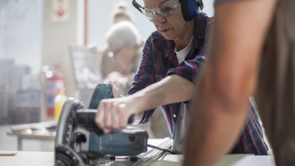 An older woman uses a power saw on some wood.