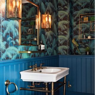 green patterned palm lead wallpaper with a gold mirror and two wall lights and a white basin on blue panelling