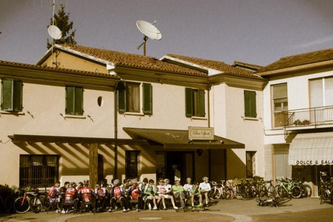 Groups of riders wait outside of a cafe as other teams are introduced.