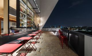 View of the terrace of the restaurant with red tables and chairs, with a skyline view of the city, at night.