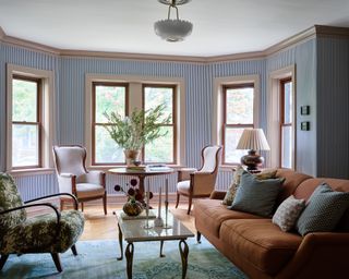 Living room with brown-orange sofa facing armchair across coffee table with rug below, striped wallpaper and bay window area with circular table and armchairs