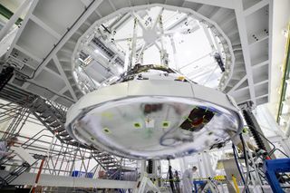 The reflective chrome bulging circular disk that is Orion's heat shield hangs below a circular scaffolding. White panels circle the edge of the heat shield where it attaches to the Orion capsule. Three technicians in masks and white coats are concentrated on different tasks.