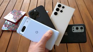 Best smartphones: Google Pixel 7a, Apple iPhone 14 Pro, Samsung Galaxy S23 Ultra, and OnePlus 10 Pro fanned out in a hand over a slatted wooden bench