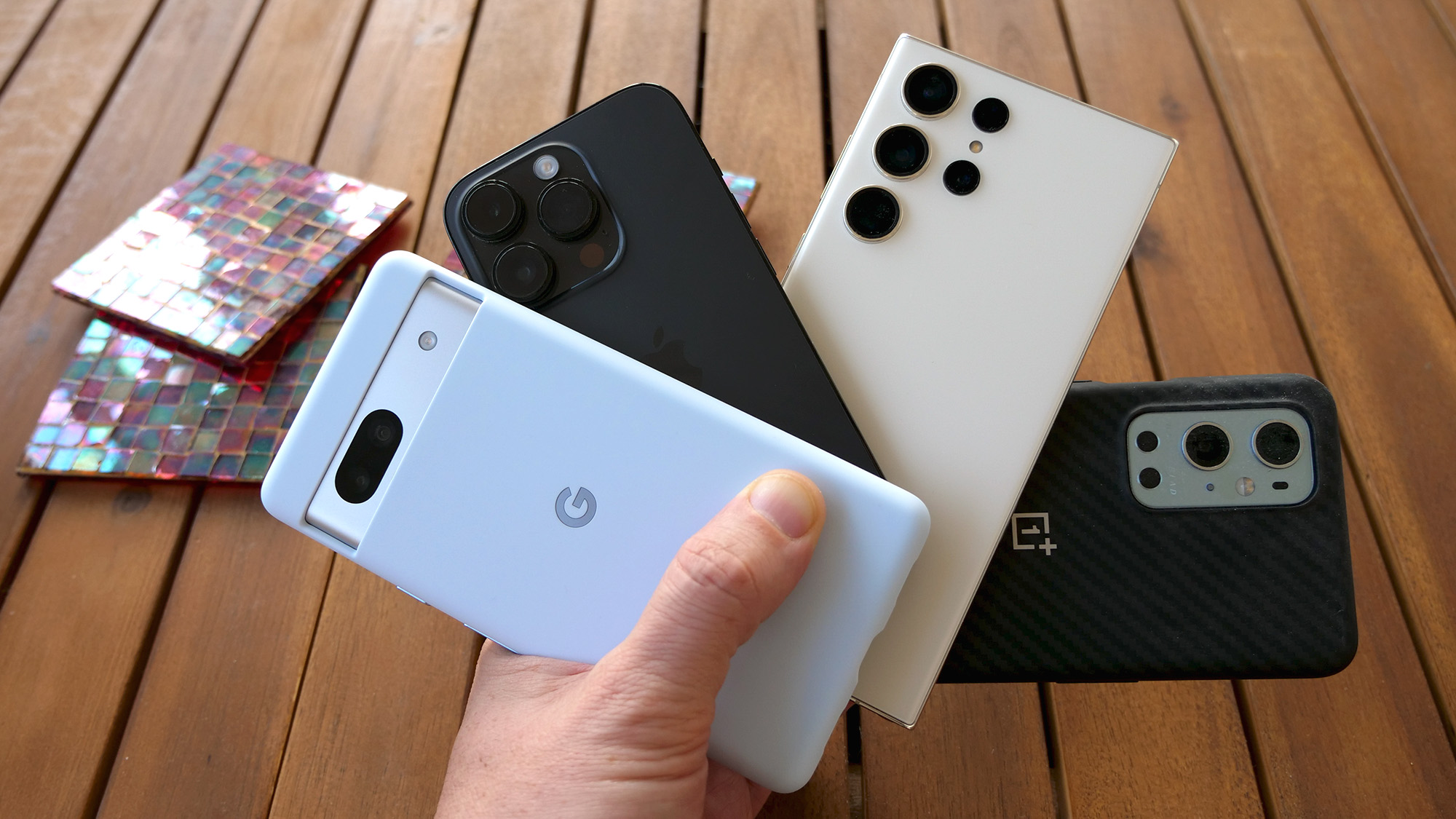 Smartphone terbaik: Google Pixel 7a, Apple iPhone 14 Pro, Samsung Galaxy S23 Ultra, dan OnePlus 10 Pro mengipasi di atas bangku kayu berpalang
