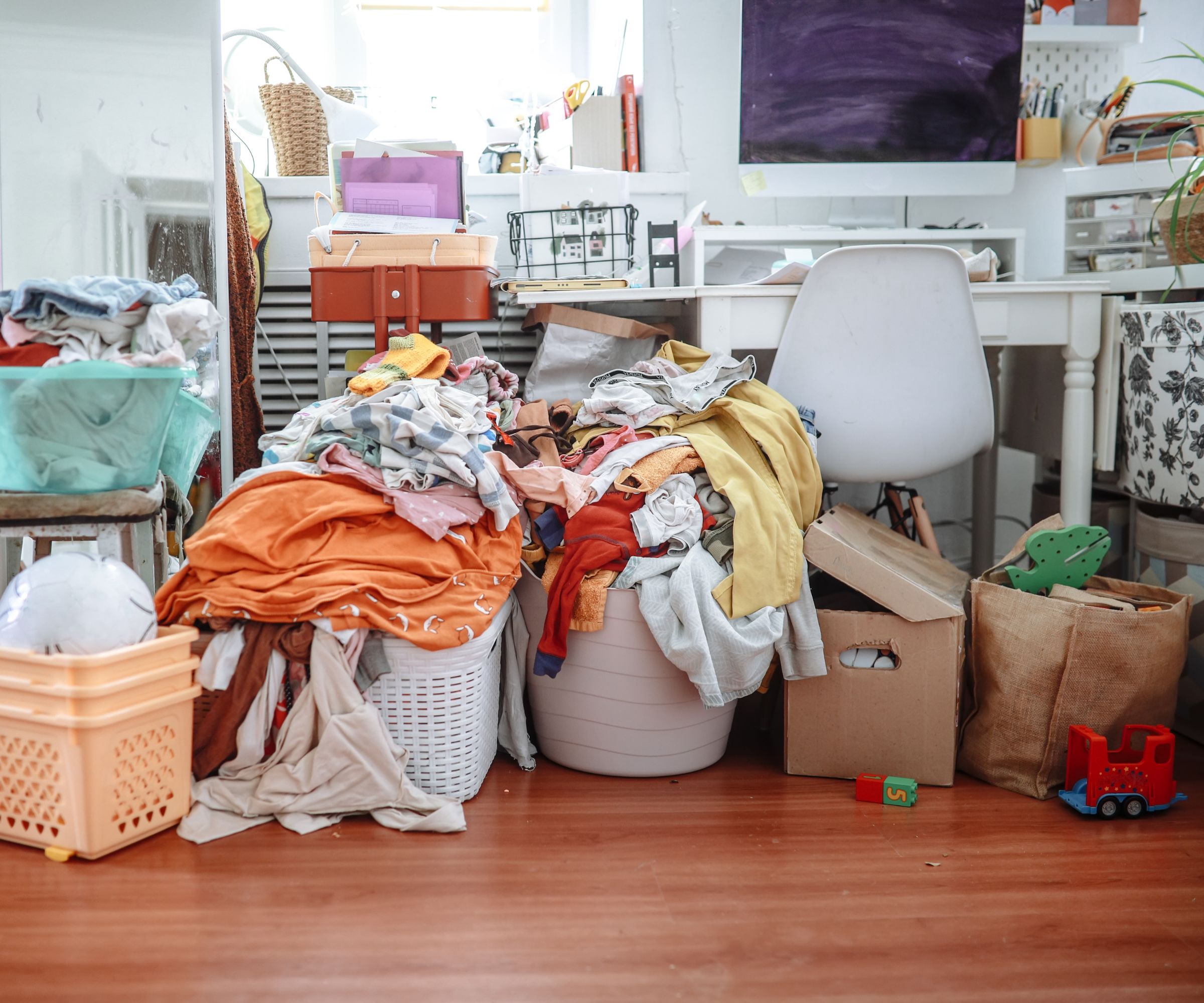 A very cluttered room with large piles of mixed clothing spilling from laundry baskets, cardboard boxes, toys and storage containers covering the floor.