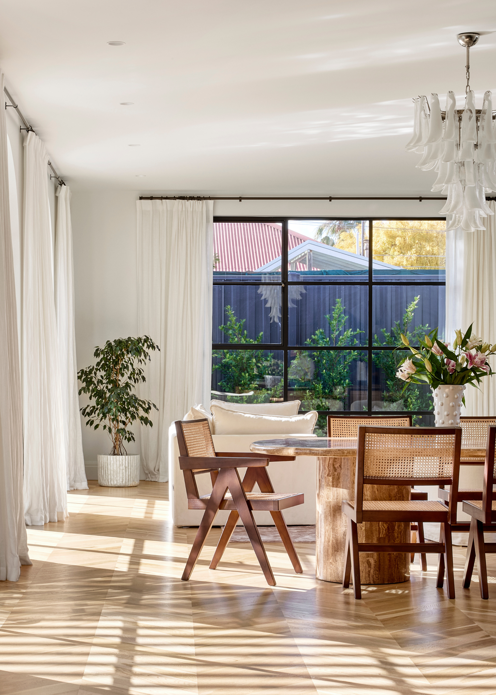 A dining room with large windows, a small potted tree, a rattan dining set, a chandelier and a vase of flowers