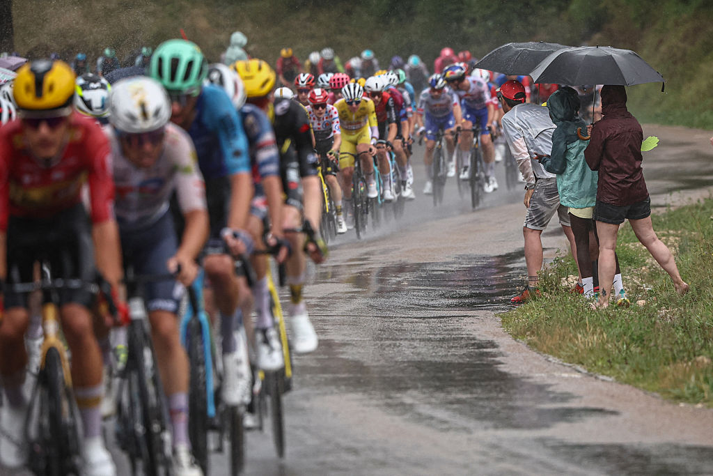 Spectators shelter from the rain under umbrellas as the pack of riders (peloton) cycles past during the 20th stage of the 112th edition of the Tour de France cycling race, 184.2 km between Nantua and Pontarlier, in the Jura, eastern France, on July 26, 2025. (Photo by Anne-Christine POUJOULAT / AFP)