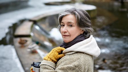 An older woman looks at the camera. She is outdoors and dressed for cold weather, holding a hot drink.