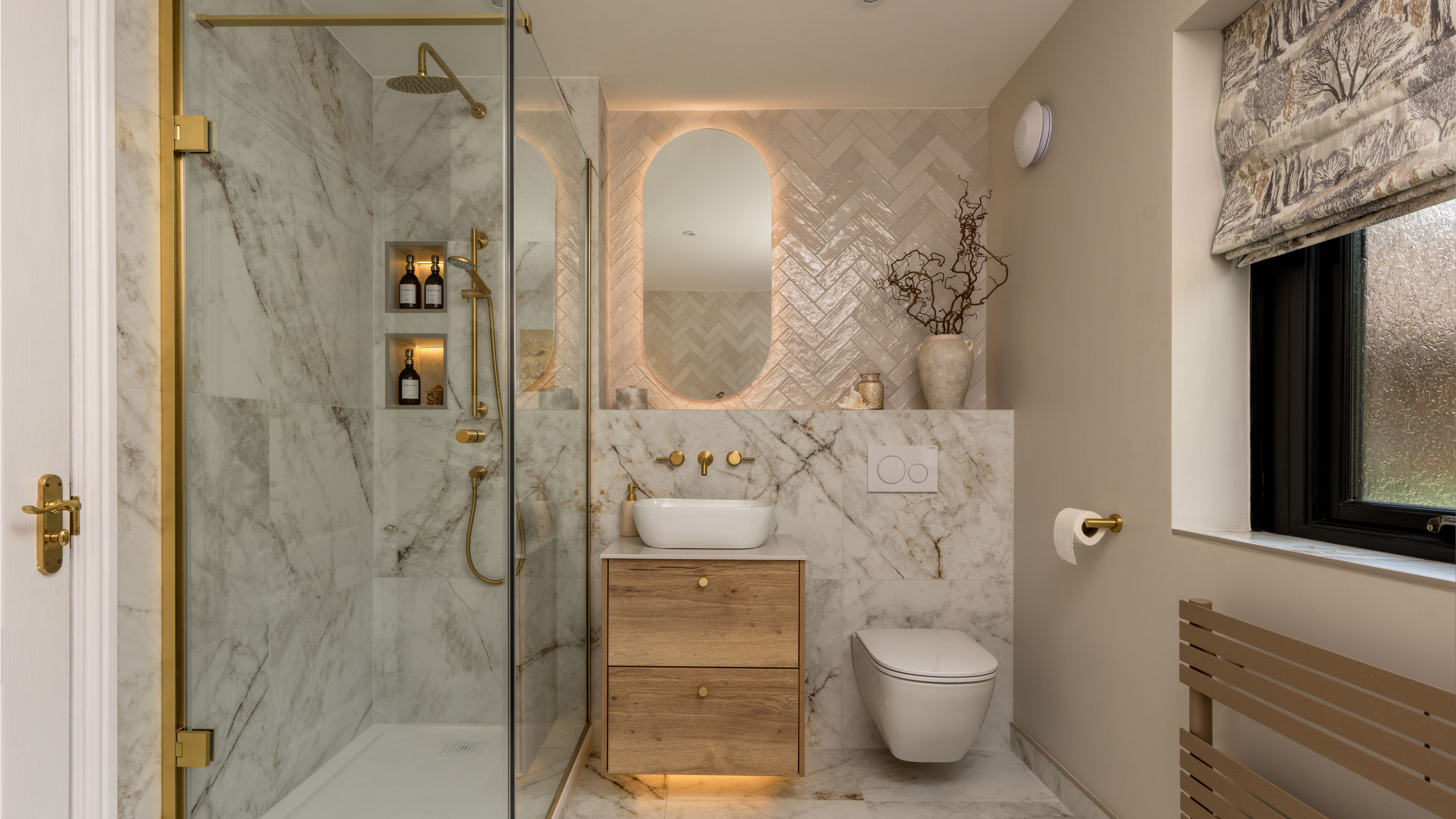 Marble shower enclosure, wooden sink vanity, herringbone tiles behind oval mirror and wall hung white bathroom.
