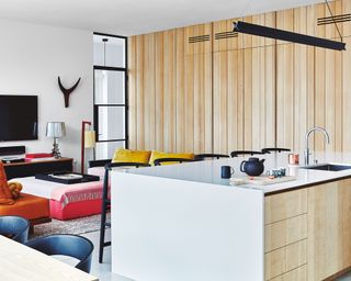 Modern open-plan kitchen with wooden cladded wall, white kitchen island, large pendant light, seating area in background