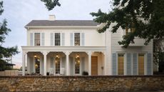 a large white Southern style home with large porch, pale blue shutters, grey roof, trees, and a stone garden wall