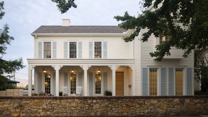 a large white Southern style home with large porch, pale blue shutters, grey roof, trees, and a stone garden wall