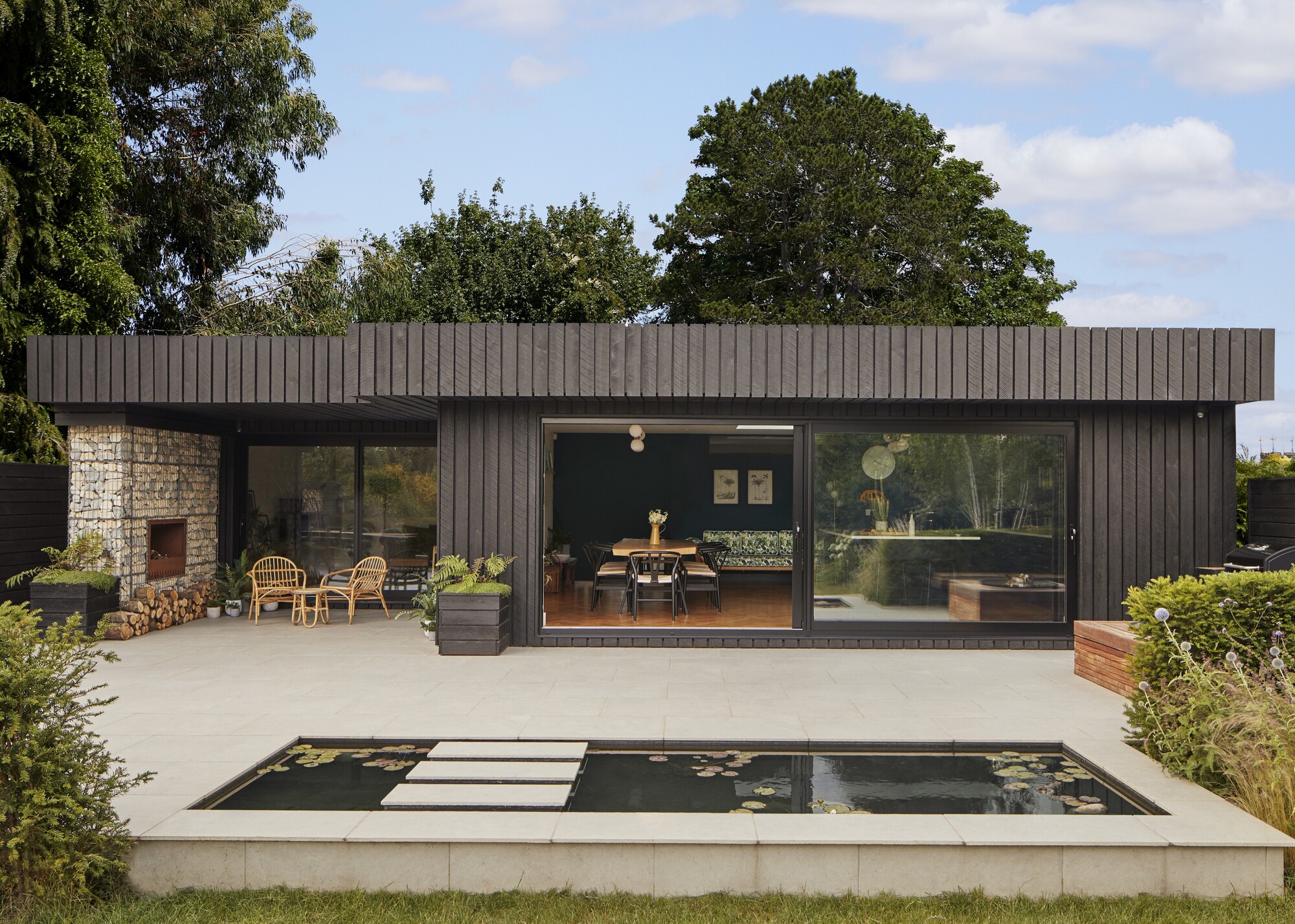 a garden room clad in black timber with large windows. There is also a view of the inside of the home with a large dining table and chairs inside.