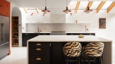 A bright white kitchen with an island, an orange accent wall with a fridge, tiger print chairs, and a skylight