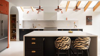 A bright white kitchen with an island, an orange accent wall with a fridge, tiger print chairs, and a skylight