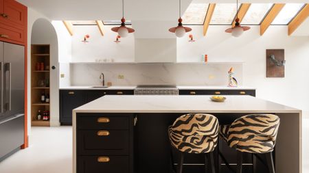 A bright white kitchen with an island, an orange accent wall with a fridge, tiger print chairs, and a skylight