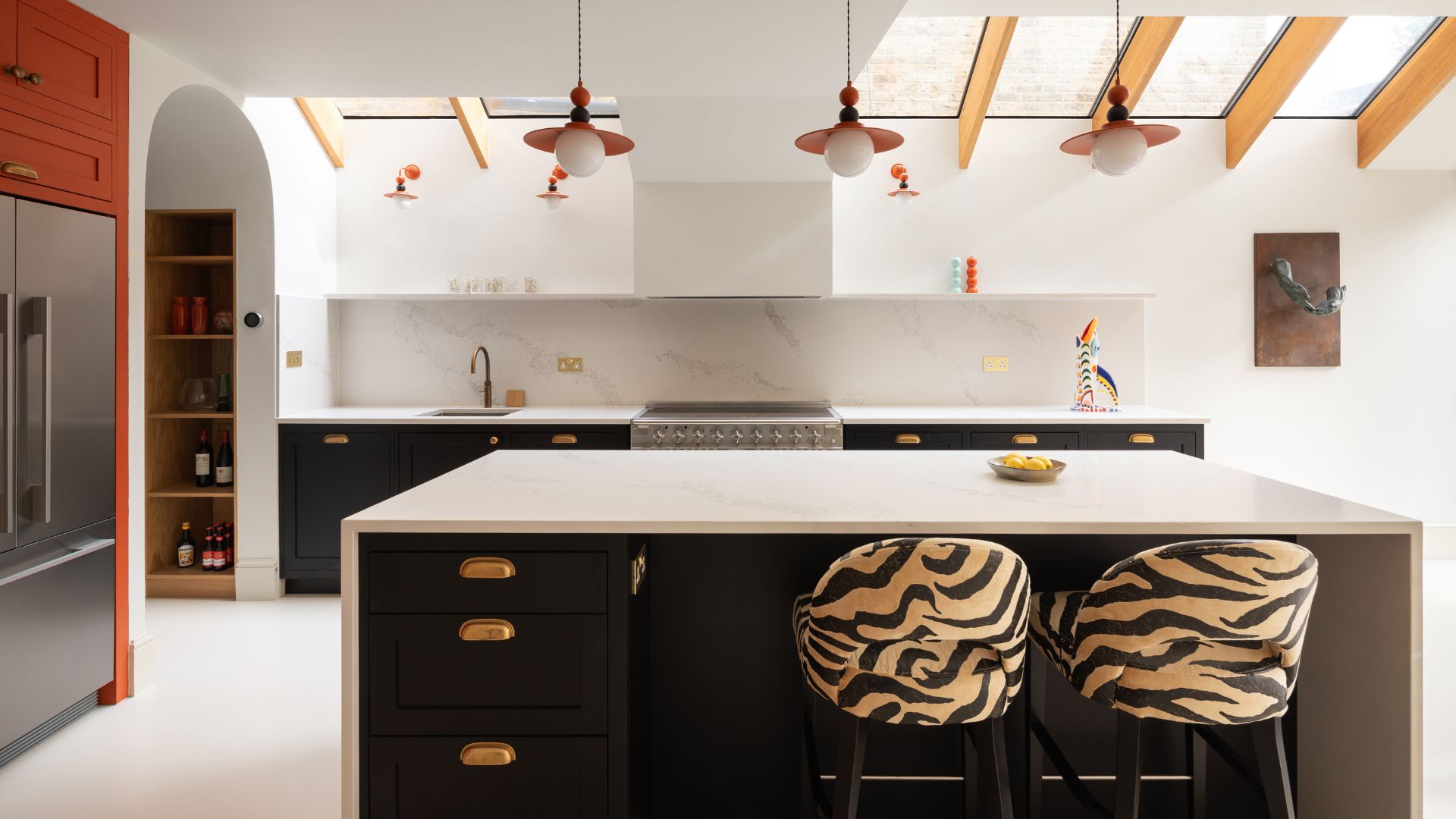 A bright white kitchen with an island, an orange accent wall with a fridge, tiger print chairs, and a skylight